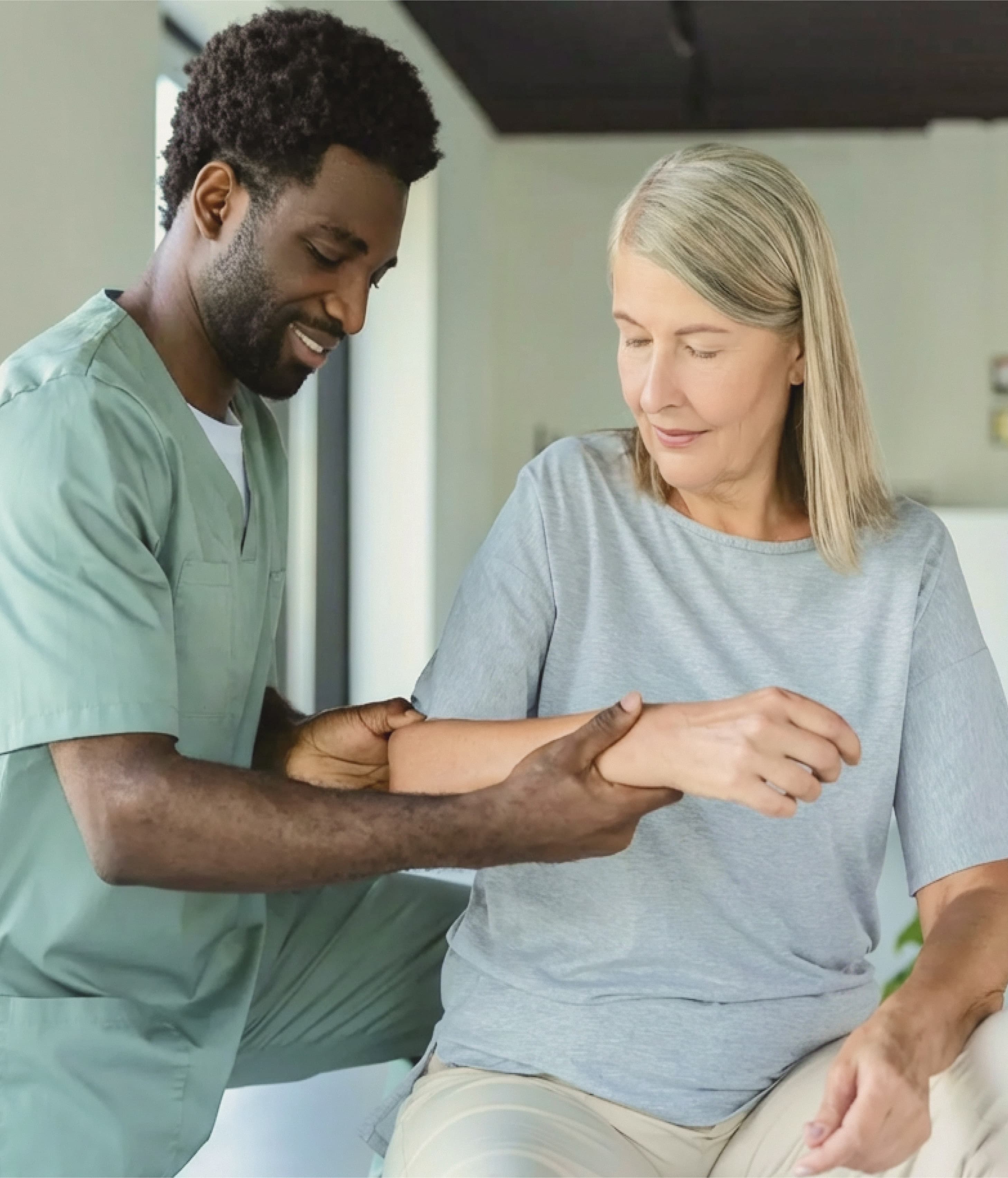 A caregiver in green scrubs gently supports an older woman’s arm. The setting is calm, conveying care and assistance in a bright room.