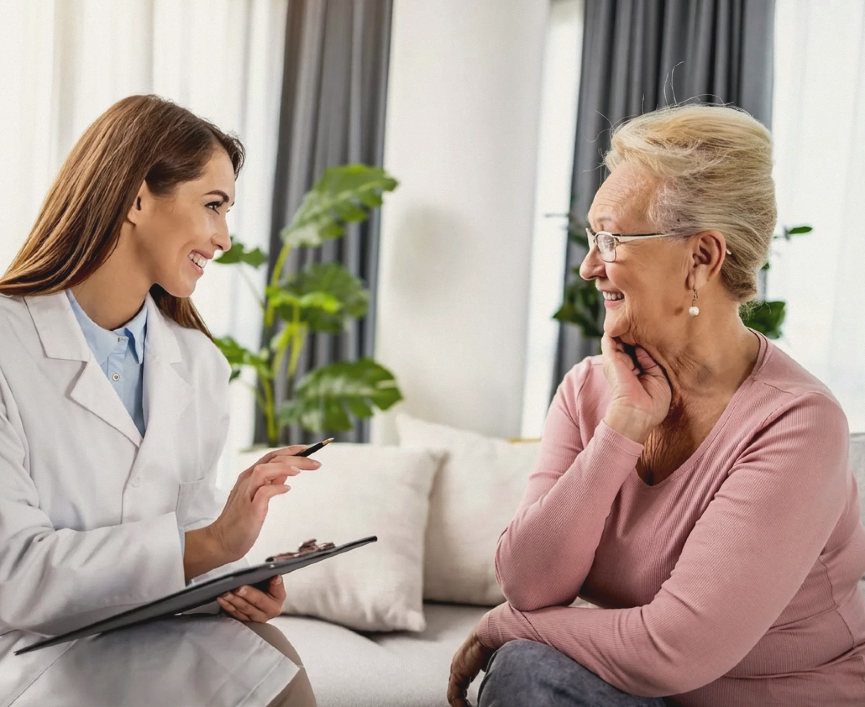 A female doctor and an elderly woman sit smiling in a cozy room with plants. The doctor holds a clipboard, creating a warm, friendly atmosphere.