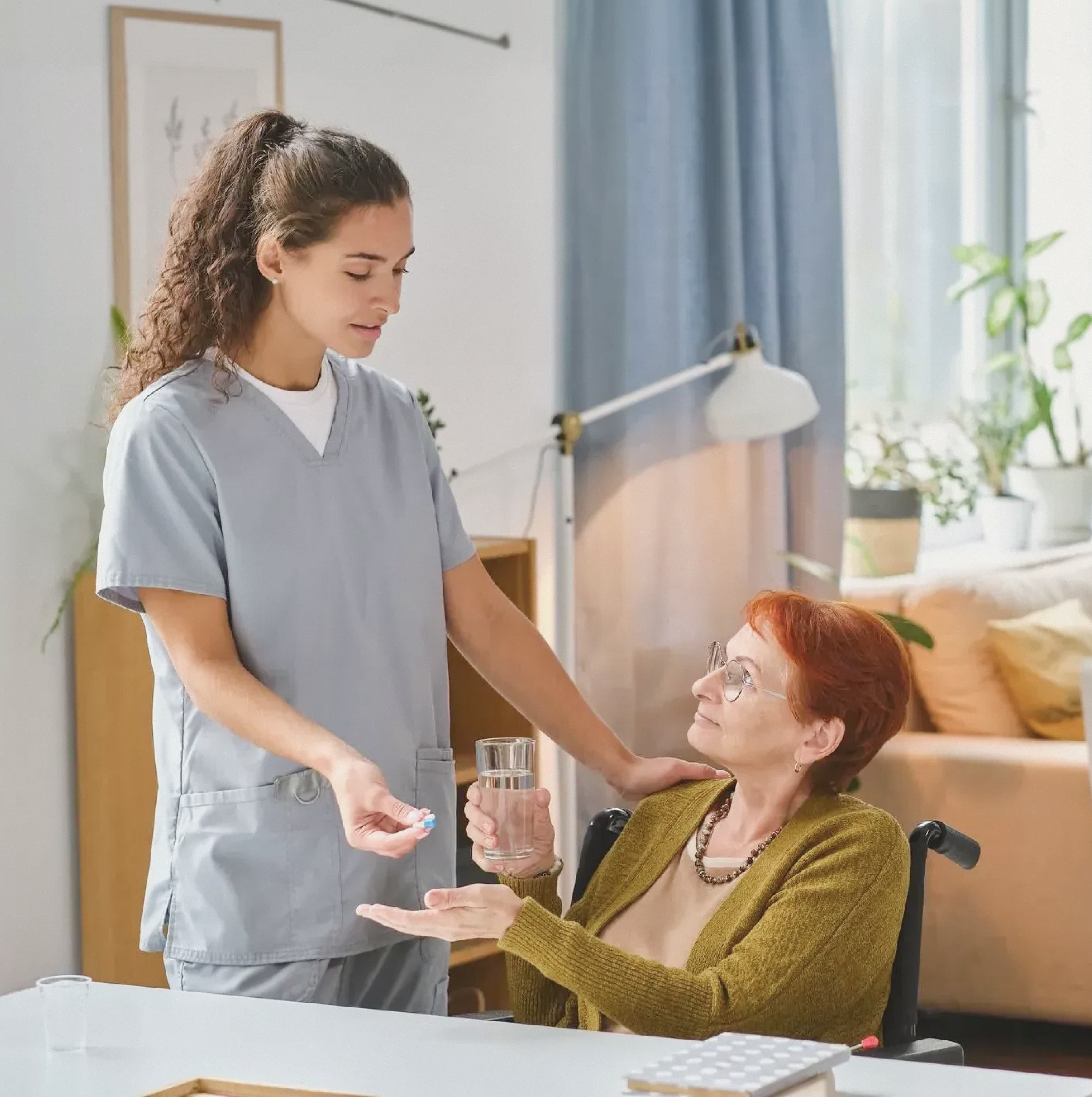 Home care nurse providing medication to elderly woman in a wheelchair at home.