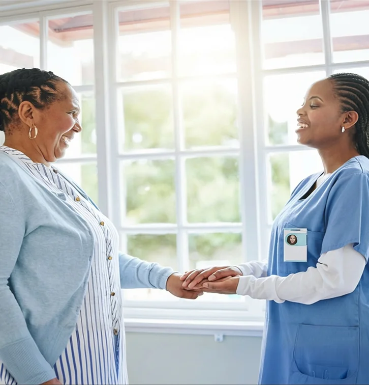 A caregiver and elderly woman holding hands and smiling warmly by a sunny window