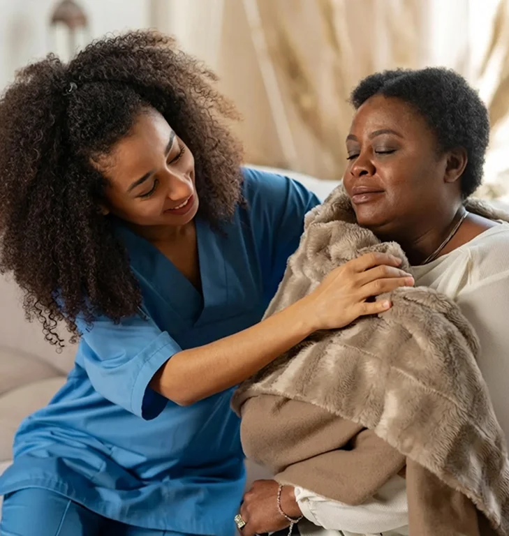 A caregiver gently wrapping a blanket around a resting elderly woman at home