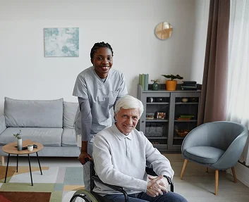 A caregiver smiling while pushing an elderly man in a wheelchair indoors