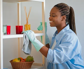 A smiling home aide dusting a shelf while wearing cleaning gloves