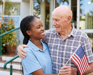 A caregiver and elderly veteran man holding an American flag smiling outdoors