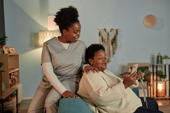 African American elderly woman learning how to use online payment services on mobile phone with help of cheerful female social worker, while sitting on couch in cozy living room