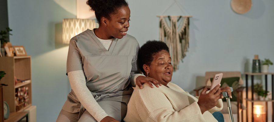 African American elderly woman learning how to use online payment services on mobile phone with help of cheerful female social worker, while sitting on couch in cozy living room