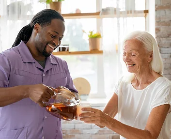 Caregiver pouring tea for smiling elderly woman in kitchen