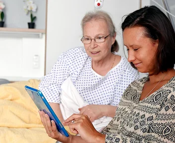 Visitor showing tablet to elderly woman in hospital bed