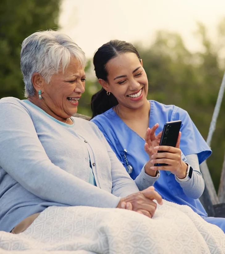 Caregiver sharing phone with laughing senior woman outdoors