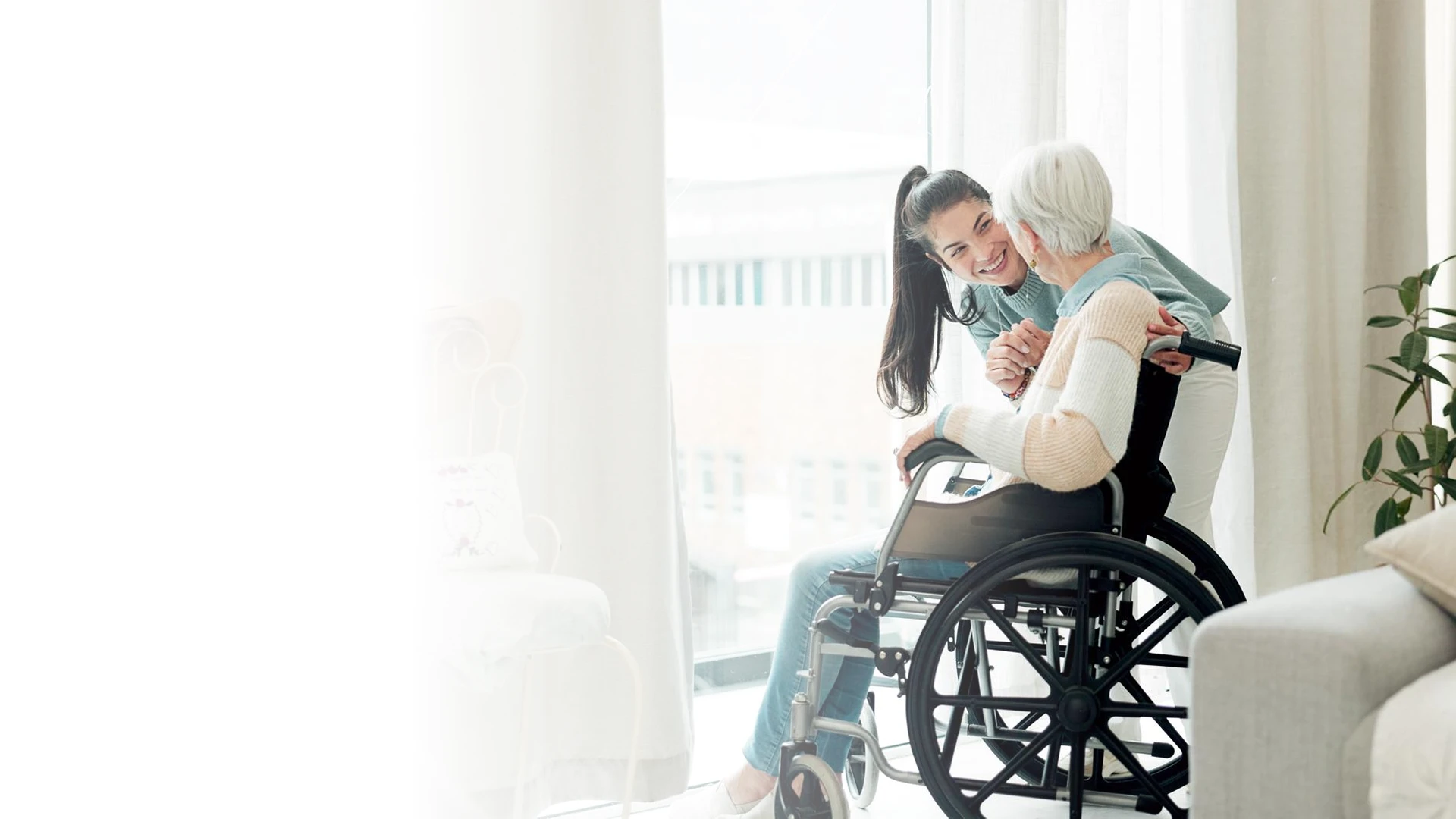 Smiling caregiver leaning in to embrace a senior woman in a wheelchair by a bright window