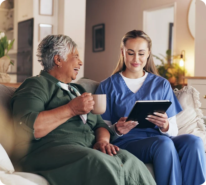 Caregiver showing a tablet to a laughing senior woman holding a mug on a sofa