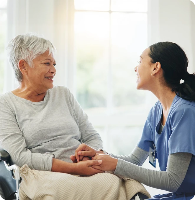Caregiver holding hands with a smiling senior woman in a wheelchair by a bright window