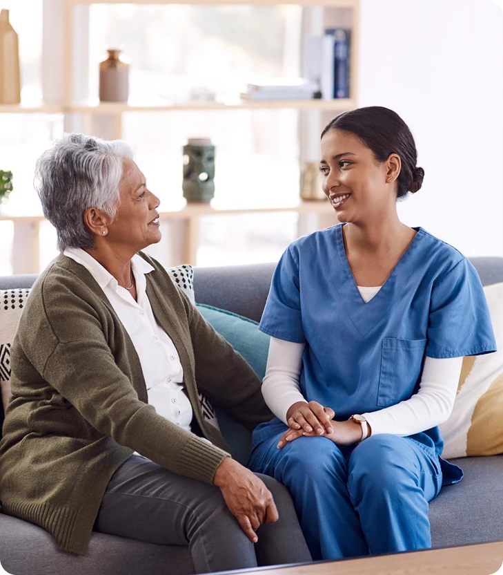 Smiling caregiver in blue scrubs talking with a senior woman on a sofa