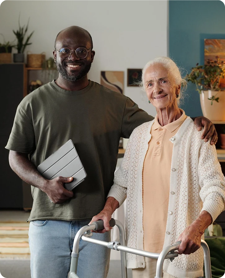 Smiling caregiver holding a tablet standing beside a senior woman with a walker at home