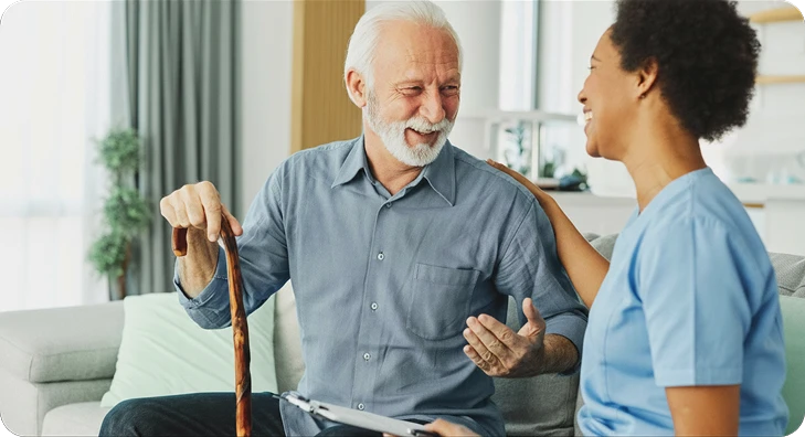 Caregiver with a clipboard having a cheerful conversation with a senior man holding a cane on a sofa