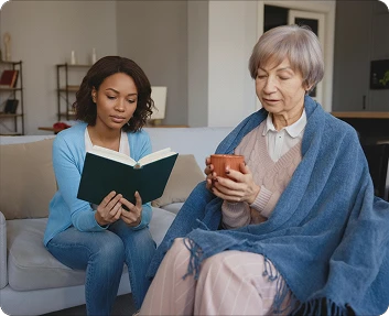 Claude responded: Caregiver reading a book aloud to a senior woman holding a mug on a sofaCaregiver reading a book aloud to a senior woman holding a mug on a sofa