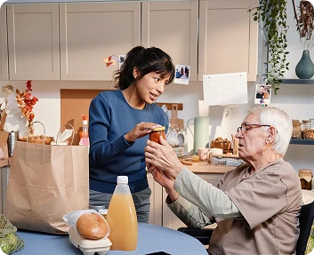 Caregiver helping a senior man open a jar while unpacking groceries in the kitchen