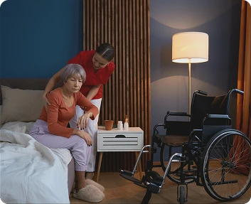 Caregiver assisting a senior woman sitting on the edge of a bed at night with a wheelchair nearby