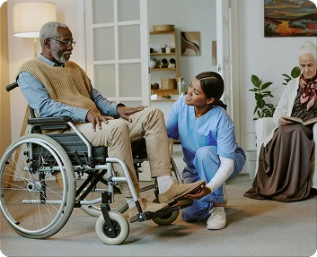 Caregiver kneeling to assist a senior man in a wheelchair while another elderly woman sits nearby