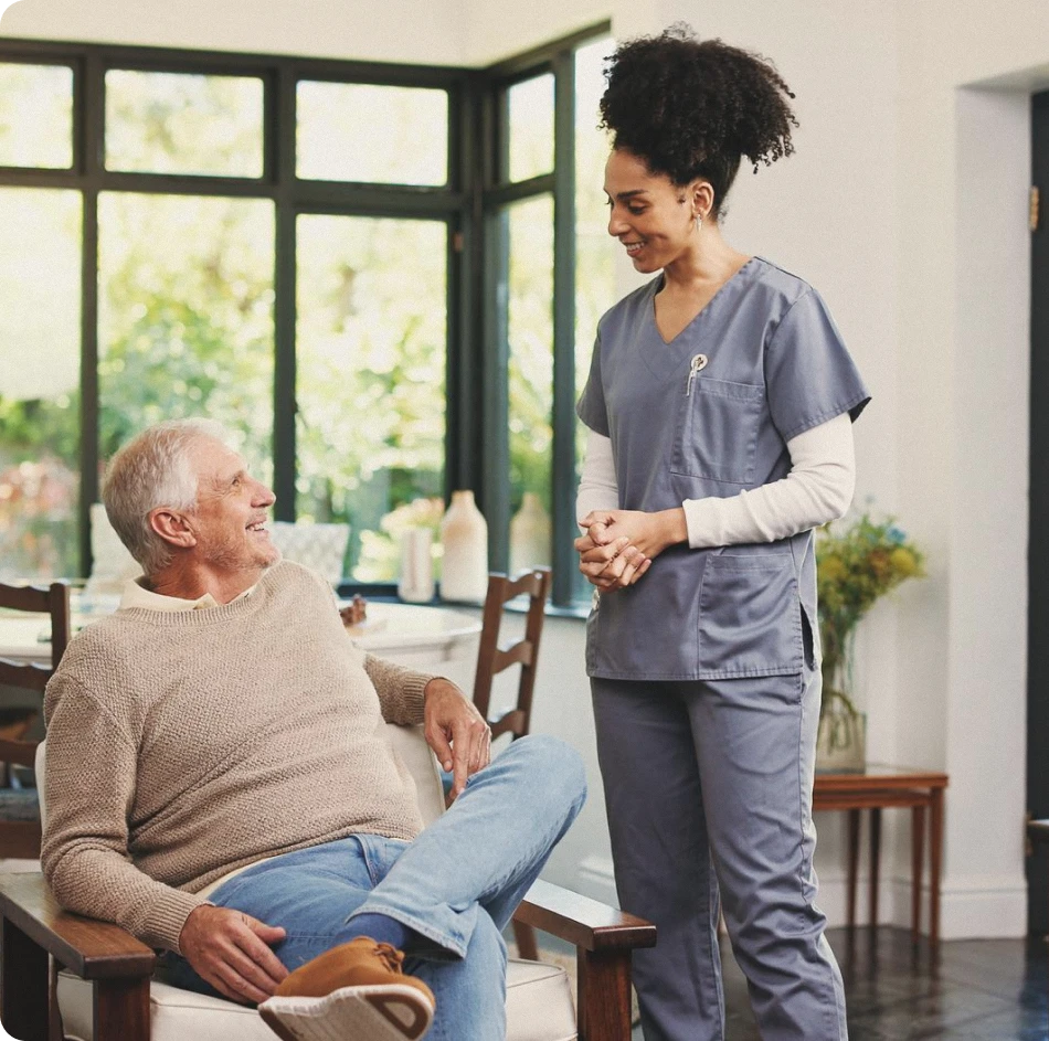Smiling caregiver in gray scrubs chatting with a senior man seated in an armchair
