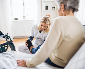caregiver checking elderly woman's leg while she sits on bed